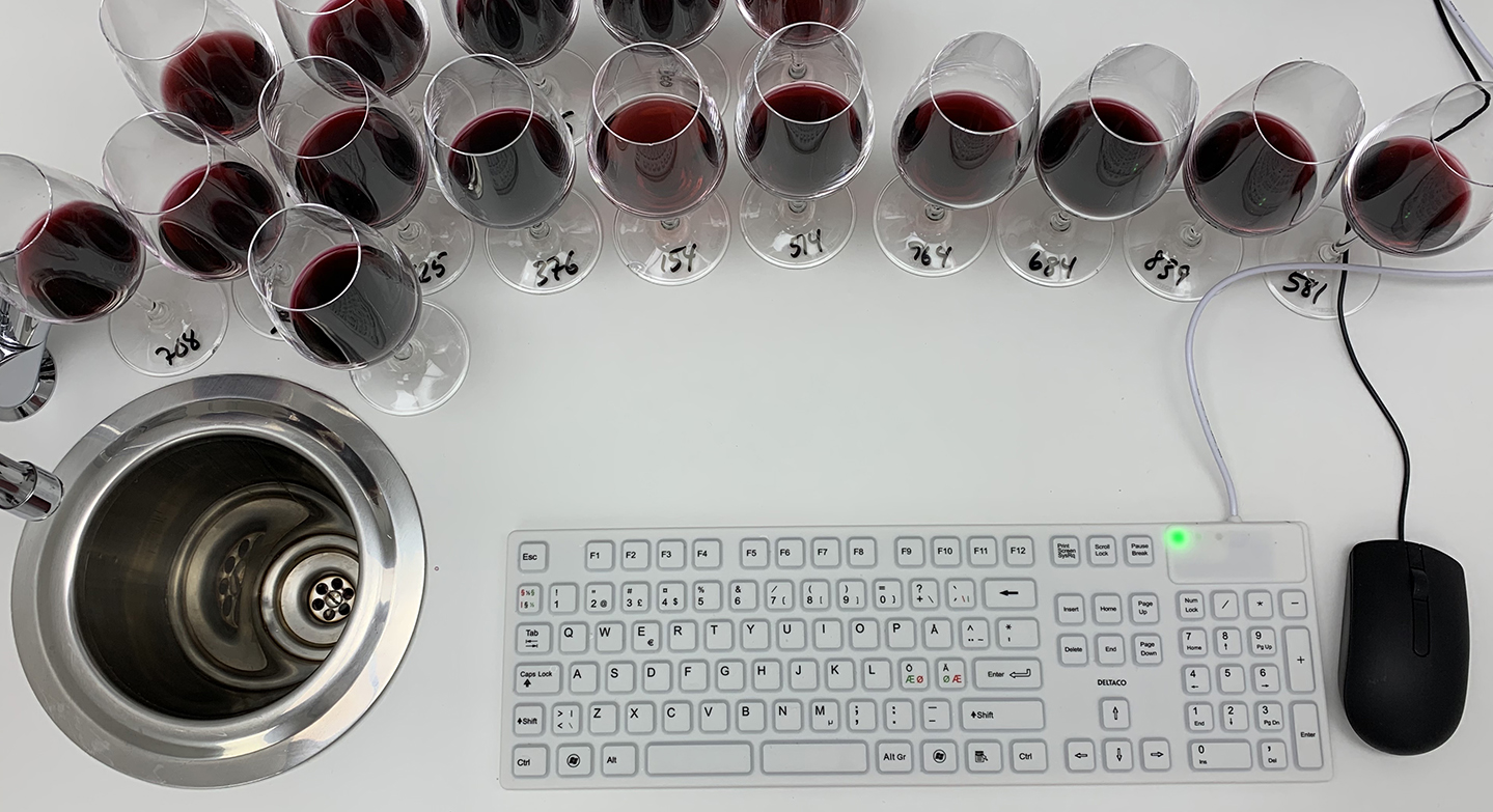 Skrivebord med to rekker rødvinsvinglass med tall på stettene, vask, mus og tastatur.