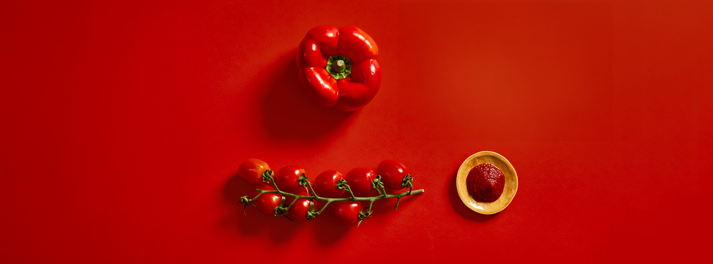rød paprika, ketchup i en skål, og en klase røde tomater på rød bakgrunn.
