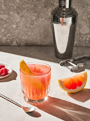 Et glass med rosa-oransje drikke, en grapefruktbåt og en drinkshaker.