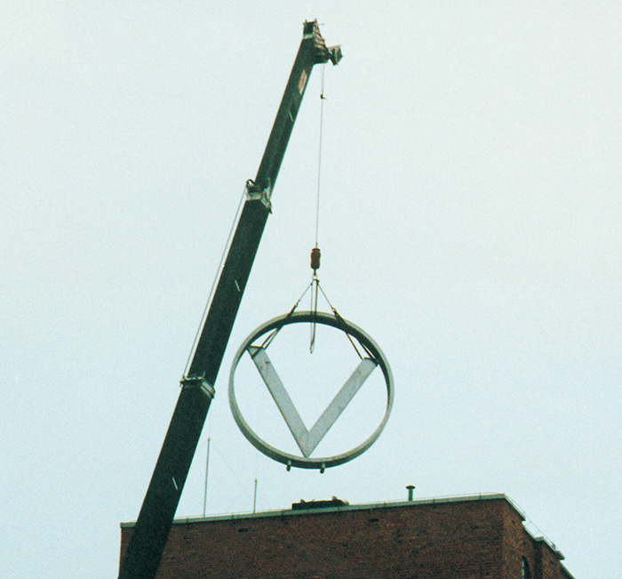 Vinmonopolets logo heises ned fra toppen av en bygning ved hjelp av en heisekran.