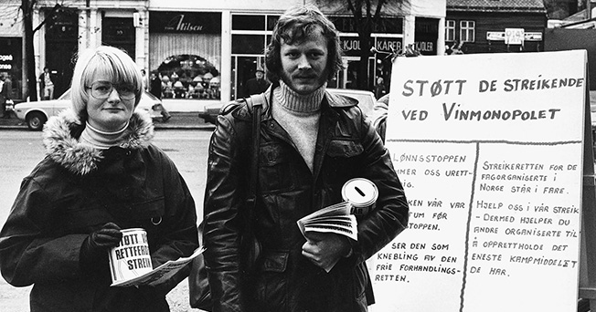 En mann og en dame med flyveblader foran en plakat med skriften &quot;Støtt de streikende ved Vinmonopolet&quot;.