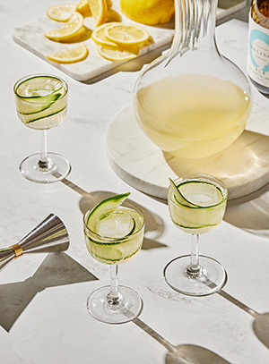 Tre stettglass lys drikke og en strimmel agurk, vinkaraffel i bakgrunnen.