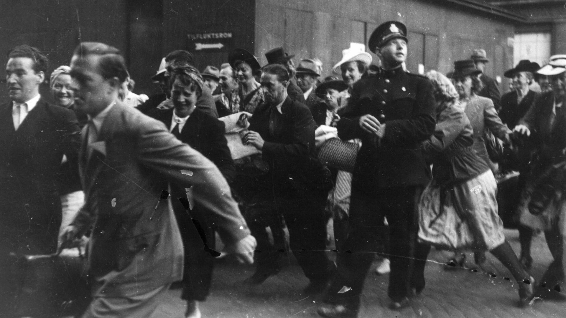 En politimann står stille blant en gruppe mennesker som løper mot venstre. Sort-hvitt bilde.