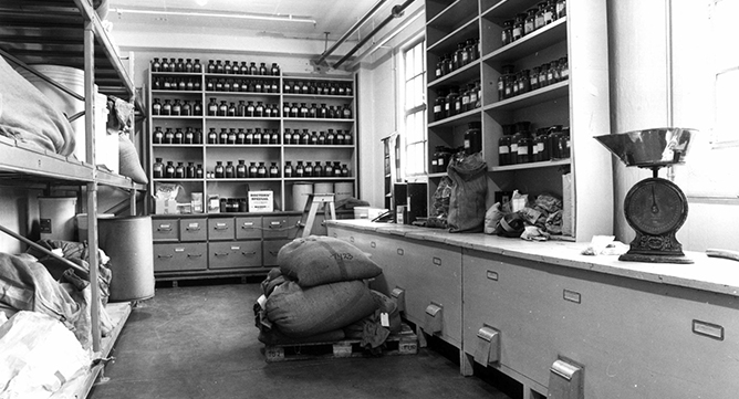 sort-hvitt bilde av interiør med hyller fulle av flasker og strieposer, og skuffer til høyre i bildet.