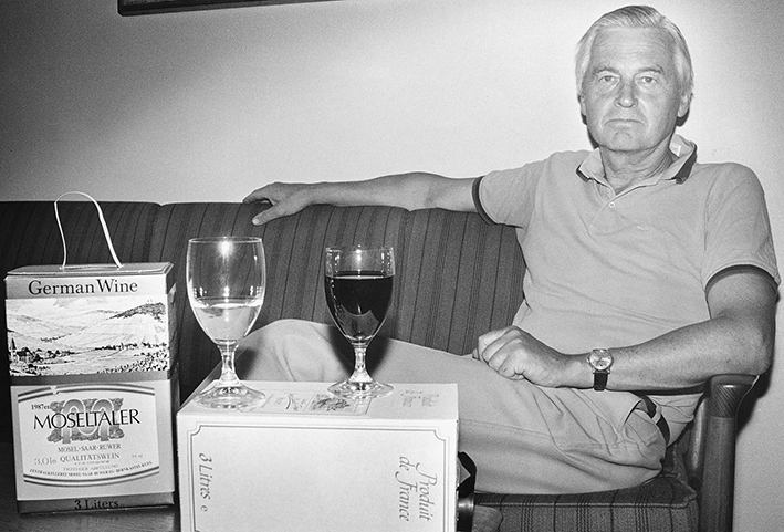 Mann sitter i en sofa. På bordet foran ham står det to vinkartonger og to glass vin.
