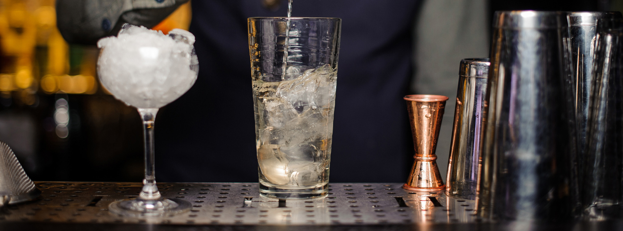 bardisk med glass, målebeger og shaker og en bartender som rører i en drink.