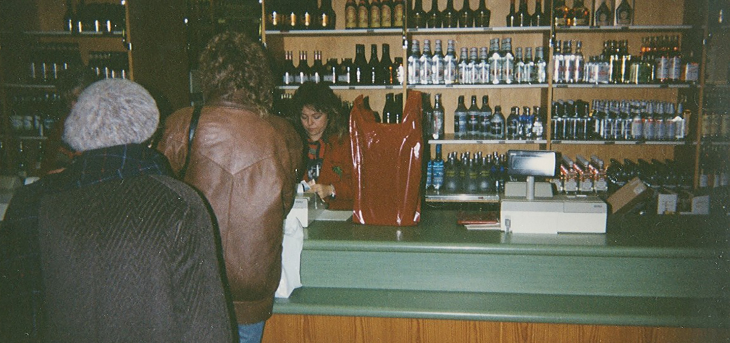 To kunder i kø foran disken der en dame betjener kundene. Bak henne er det hyller med flasker.