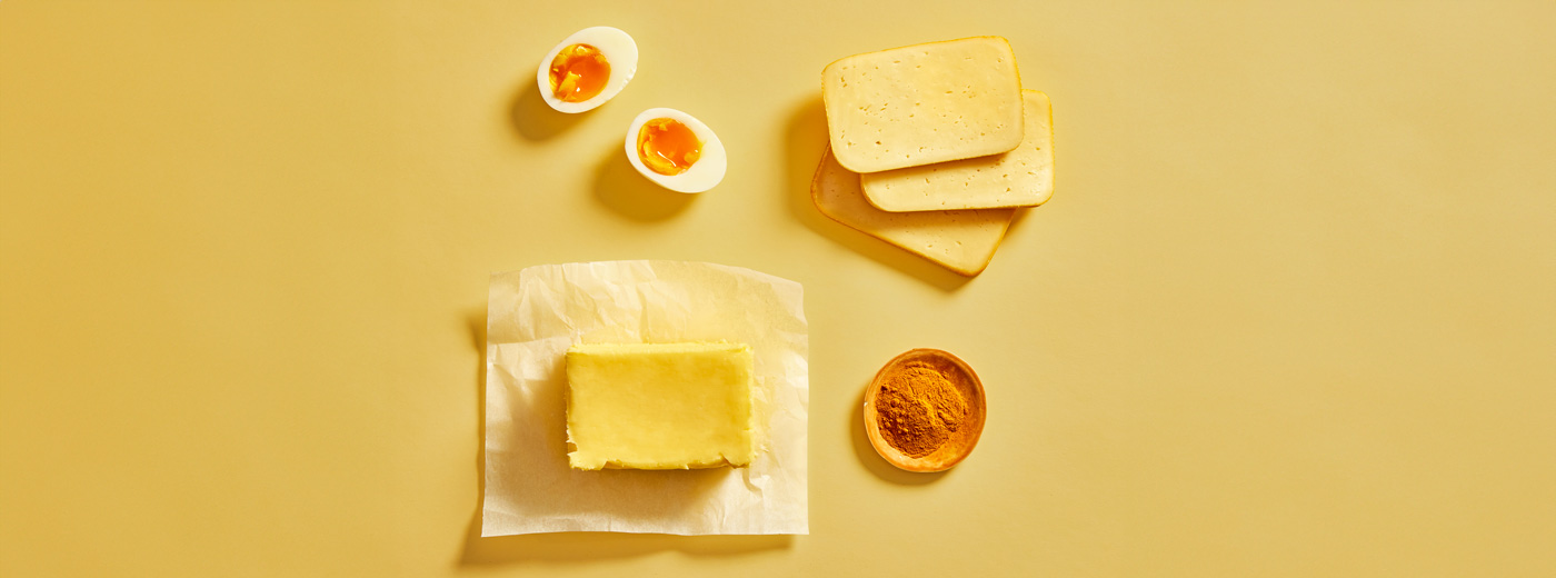 Osteskiver, en skål med gurkemeie, hardkokt egg delt i to og en pakke smør på gul bakgrunn.