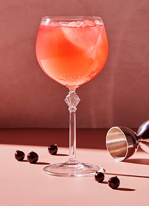 Høyt stettglass med rosa drikke og solbær på bordet ved siden av.