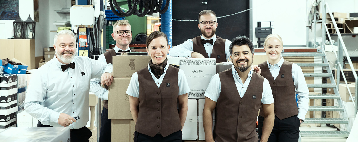 Seks ansatte med uniform står på et lager og smiler mot kamera.