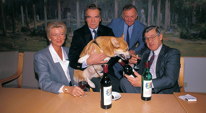 En dame til venstre og tre menn, alle i dressjakker. En av dem holder en hund på fanget, og på bordet foran dem står det tre flasker.